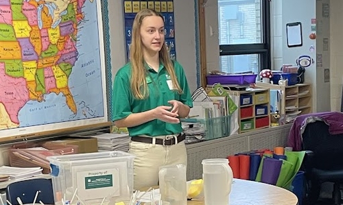 A teen girl wearing a green 4-H polo talking in front of a classroom.